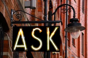 photo of metal sign with the word "ASK" in bright neon letters, hanging on a building
